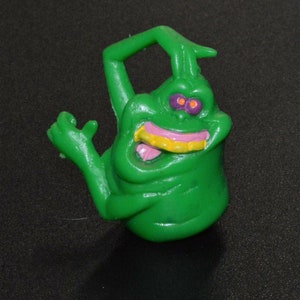Op de afbeelding: Groene plastic geestfiguur met een tandenrij, paarse ogen en een gele tong. De geest heeft een opgeheven arm en staat in een speelse pose.