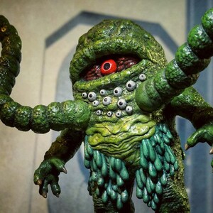 Op de afbeelding: Een groen, meerogig monster met &eacute;&eacute;n groot rood oog. Het monster heeft meerdere tentakels en een getextureerd, slijmerig lichaam.