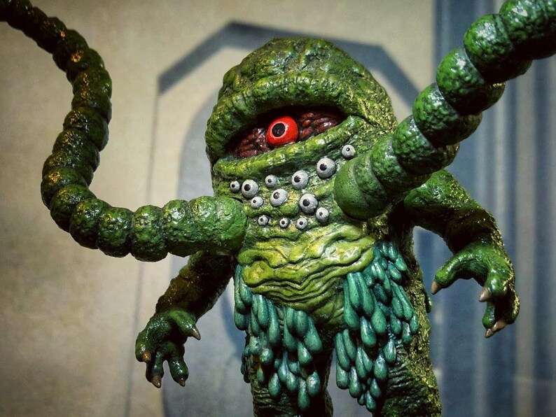 Op de afbeelding: Een groen, meerogig monster met &eacute;&eacute;n groot rood oog. Het monster heeft meerdere tentakels en een getextureerd, slijmerig lichaam.