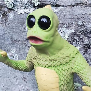 May include: A green, lizard-like figurine with large, black eyes and a yellow belly. The creature has a textured, scaly appearance and is posed with an outstretched arm. The background is a textured, gray stone surface.