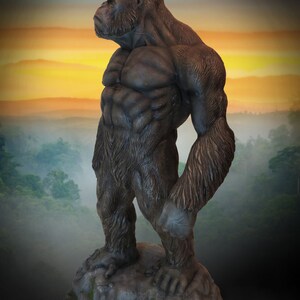 May include: A large, dark grey gorilla statue with a muscular build, standing on a rocky base. The gorilla is looking to the left, with its head tilted slightly. The statue is set against a backdrop of a foggy forest and a sunset sky.