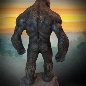 May include: A large, dark brown gorilla statue standing on a rocky outcrop. The gorilla is facing away from the viewer, with its back and muscular arms visible. The statue is set against a backdrop of a foggy forest and a colourful sunset.