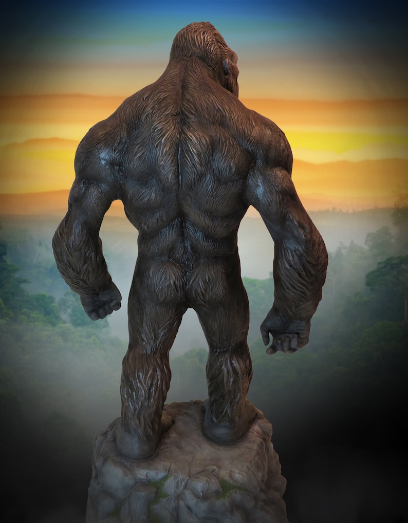 May include: A large, dark brown gorilla statue standing on a rocky outcrop. The gorilla is facing away from the viewer, with its back and muscular arms visible. The statue is set against a backdrop of a foggy forest and a colourful sunset.