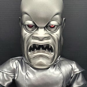 Peut inclure: Gros plan sur une poupée monstre argentée avec une expression de colère. La poupée a des yeux rouges, une bouche grande ouverte avec des dents pointues et un éclat métallique. La poupée porte une tenue argentée.