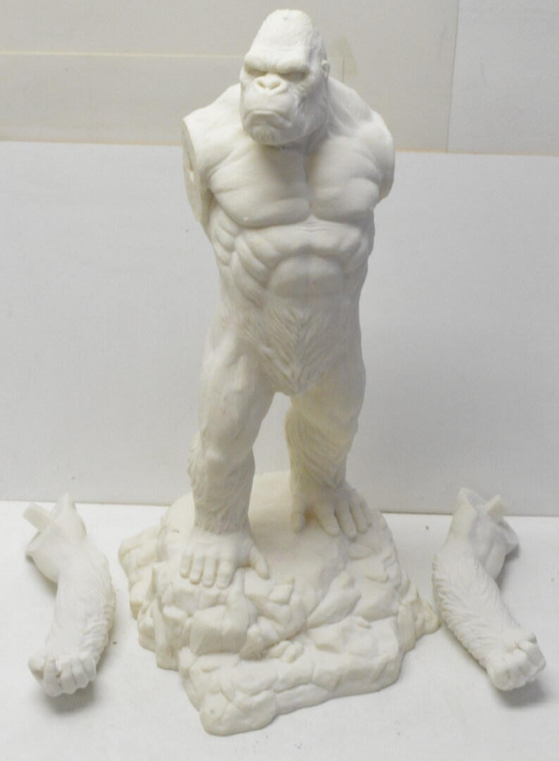 May include: An unpainted, plastic model kit of a gorilla standing on a rocky base. The model is in pieces, with two arms detached.