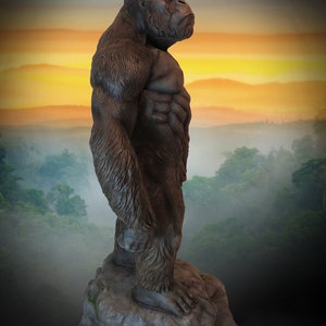 May include: A large, dark brown gorilla statue standing on a grey rock. The gorilla is facing to the right and has a muscular build. The statue is set against a backdrop of a foggy forest and a sunset sky.