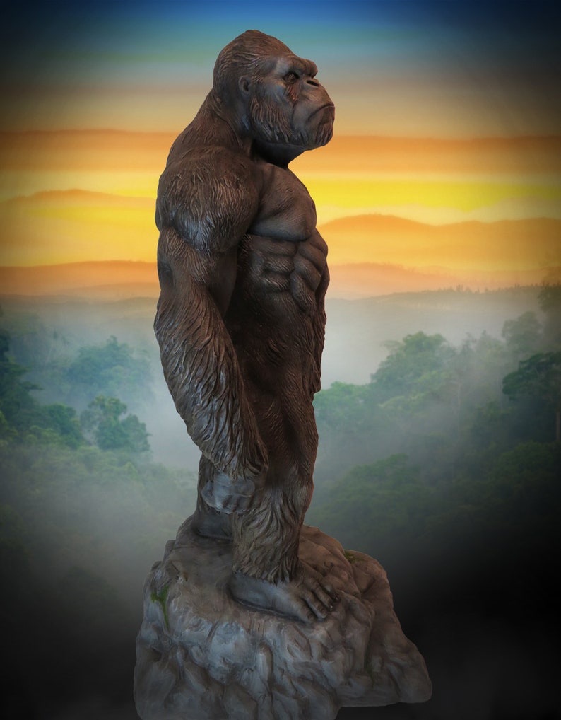 May include: A large, dark brown gorilla statue standing on a grey rock. The gorilla is facing to the right and has a muscular build. The statue is set against a backdrop of a foggy forest and a sunset sky.