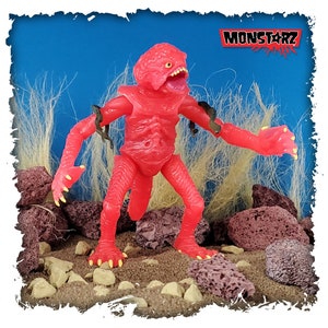 May include: A bright red, articulated action figure of a monster with a large mouth and four arms. The figure is posed in a walking position with its arms outstretched. The figure is standing on a brown, sandy surface with rocks and grass in the background. The figure is labelled "MONSTARZ" in the top right corner of the image.