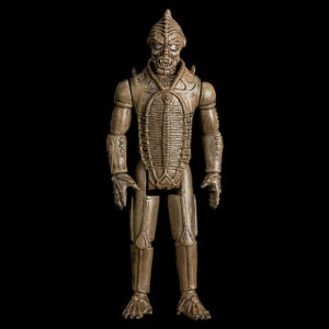 Puede incluir: Figura de acción articulada de un alienígena humanoide con un acabado texturizado marrón-beige. La figura tiene una cara esquelética, cabeza puntiaguda y detalles intrincados tipo armadura en el torso y las extremidades. Sobre fondo negro.