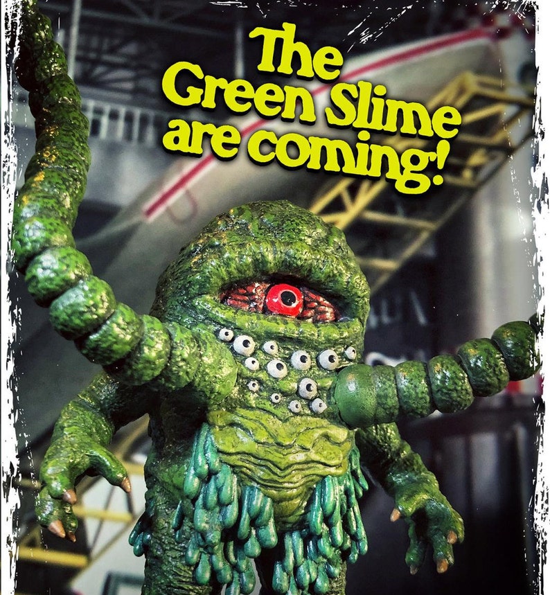 Op de afbeelding: Een groen, slijmerig monster met meerdere ogen en &eacute;&eacute;n groot rood oog. Het monster heeft een lange, groene, gesegmenteerde arm die uitstrekt. De tekst "The Green Slime are coming!" is geschreven in gele letters op een witte achtergrond.