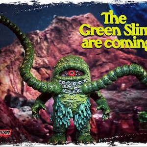 Op de afbeelding: Een groen, slijmerig monster met meerdere ogen en tentakels. Het monster staat op een rotsachtig oppervlak met een rood oog en de tekst "The Green Slime are coming!" in gele letters.
