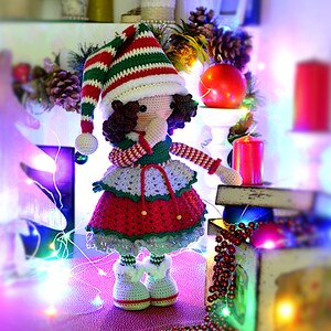 May include: A handcrafted crochet doll in an elf costume, perfect for Christmas. The doll has a red, white, and green striped hat, a red and white dress, and white boots. The scene includes Christmas lights and candles, creating a festive atmosphere.