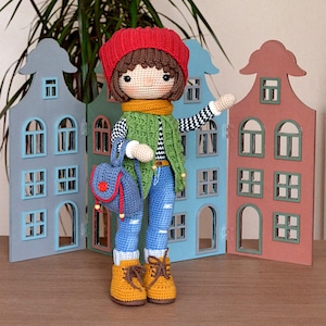 Op de afbeelding: Een gehaakte pop met een rode muts, een groene vest, een blauwe spijkerbroek en bruine laarzen. De pop houdt een blauwe rugzak vast en staat voor een achtergrond van miniatuurhouten huizen.