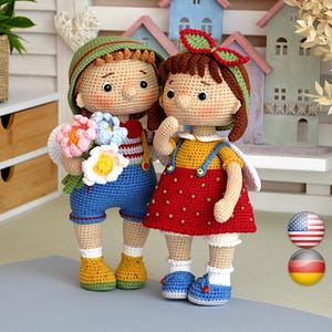 Pode incluir: Duas bonecas de crochê feitas à mão, um menino e uma menina, estão lado a lado. O boneco do menino usa um chapéu verde, macacão azul e segura um buquê de flores. A boneca da menina usa um vestido vermelho com alças amarelas e um laço vermelho.