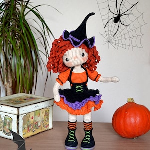 May include: A crocheted witch doll with red curly hair, wearing a black witch hat with a purple brim, an orange and black dress, and black boots with green accents. The doll is standing on a wooden surface with a pumpkin, a bat, and a spider web in the background.