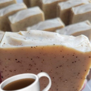 May include: A close-up of a bar of coffee-scented soap with a cup of coffee in the foreground. The soap is light brown with dark brown flecks throughout. The cup of coffee is white with a brown handle.