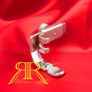 May include: Silver sewing machine foot with a screw on the top and the text "121B77 SIMANCO" on the side.