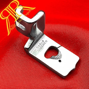 Puede incluir: Un pie de máquina de coser Simanco 88818 plateado y brillante, diseñado para coser. El pie tiene una forma curva con una base plana y una abertura en forma de corazón. El fondo es rojo liso.