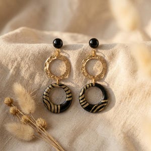 May include: A pair of drop earrings featuring a black stud, a textured gold-tone circular link, and a larger black circular pendant with gold-tone striped patterns. The earrings are constructed with a vertical arrangement of three distinct circular elements.