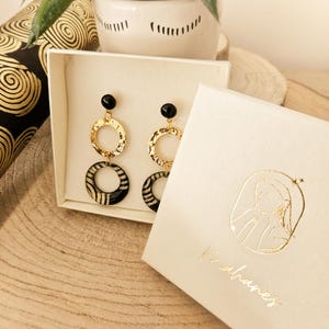 May include: A pair of drop earrings featuring a black stud, a textured gold-tone ring, and a bottom ring with a black and white striped pattern. The earrings are presented in a white gift box with a gold logo and the text Mahanès printed on it.