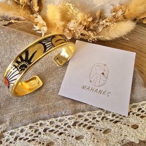 Puede incluir: Un brazalete dorado con un diseño de sol negro y amarillo. El brazalete se exhibe sobre una superficie de tonos neutros con una tarjeta que dice "MAHANÉS". También se ven flores secas y un ribete de encaje.