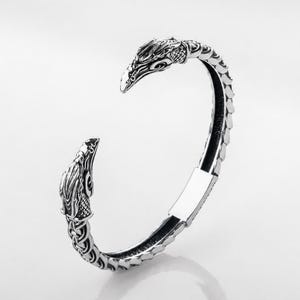 May include: A silver-toned metal cuff bracelet featuring two detailed bird head terminals at each end. The band has a textured, scale-like pattern along the outer surface, with intricate engravings on the heads and a smooth, flat section at the back.