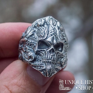 Peut inclure: Bague argent&eacute;e d&eacute;taill&eacute;e repr&eacute;sentant un cr&acirc;ne avec des gravures complexes. La bague pr&eacute;sente un design de cr&acirc;ne avec des traits faciaux d&eacute;taill&eacute;s et des motifs orn&eacute;s. La bague est tenue dans une main, avec un arri&egrave;re-plan flou.