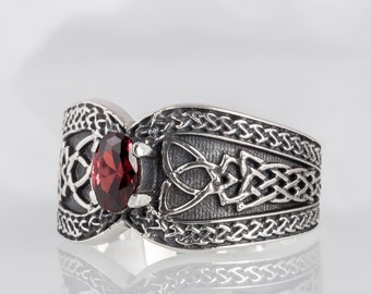 May include: A silver ring featuring an oval-cut dark red gemstone set in the center. The band is crafted with intricate, raised metalwork displaying a continuous woven knot pattern across the surface, creating a textured and detailed appearance.