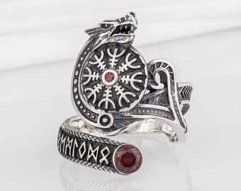 May include: Silver ring with a dragon design and a red gemstone. The ring features a circular design with a black and white pattern and the word "Vegvisir" inscribed on the band.
