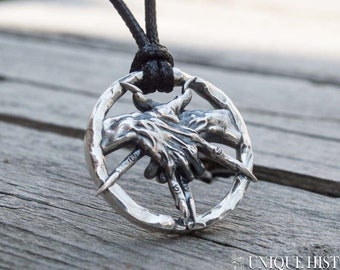 May include: A silver pendant necklace featuring a circular design with two hands clasped together, each hand holding a knife. The pendant is on a black cord.