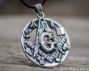 May include: Silver-toned pendant featuring a Masonic symbol, including a square, compass, and other tools, suspended from a black cord. The pendant has intricate detailing and is set against a blurred wooden background. The shop's name, "UNIQUE HISTORY SHOP", is at the bottom.