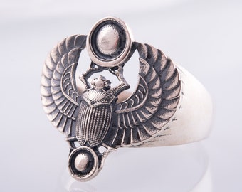 May include: A silver ring featuring a scarab beetle design. The beetle is holding a sun disc in its front legs. The wings are spread out behind the beetle.
