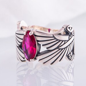 May include: A silver ring with a marquise-cut, bright pink gemstone. The ring's design includes stylised wings on either side of the gemstone, with detailed feather etching. The ring is set against a white background.