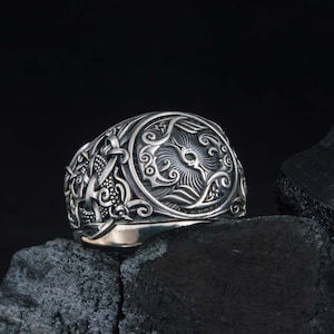 May include: A silver-colored ring with an ornate design featuring a central sunburst motif and intricate scrollwork. The ring is set against a dark background, highlighting its detailed craftsmanship. The ring appears to be made of metal.