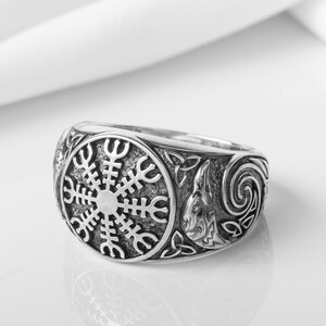 May include: A silver ring with a detailed design featuring a circular symbol and a Celtic knot. The symbol is a stylized sun wheel with eight spokes.