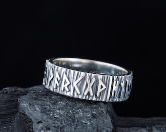 May include: A silver ring with a textured band and runes engraved around the band. The ring is resting on a piece of black charcoal.