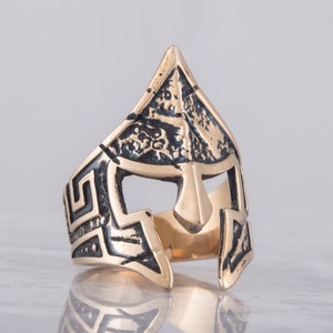 Pode incluir: Um anel dourado com um design detalhado de um capacete espartano. O capacete &eacute; texturizado e tem um padr&atilde;o de chave grega ao redor da banda.