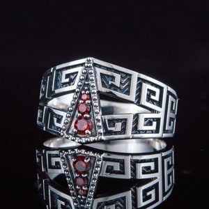 May include: A silver ring with a geometric design and red gemstones. The ring is set on a black background.