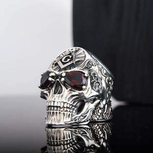 May include: A silver skull ring with red gemstone eyes and a Masonic symbol on the forehead. The ring is detailed with intricate carvings and is set against a black background.
