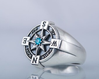 Puede incluir: Un anillo de plata con forma de brújula con una piedra preciosa azul en el centro. Los puntos cardinales de la brújula están etiquetados con los cuatro elementos: fuego, agua, tierra y aire.