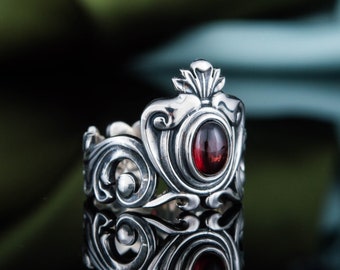 Pode incluir: Anel de prata ornamentado com um design semelhante a uma coroa. O anel apresenta uma pedra preciosa central oval, vermelha escura, rodeada por intrincados arabescos e elementos decorativos. O anel é altamente reflexivo, com fundo escuro.