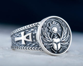 May include: A silver ring with an engraved design. The ring features an Ankh symbol on the band and a detailed circular emblem with a scarab beetle, sun, and wings. The ring has a textured finish and is set against a dark background.
