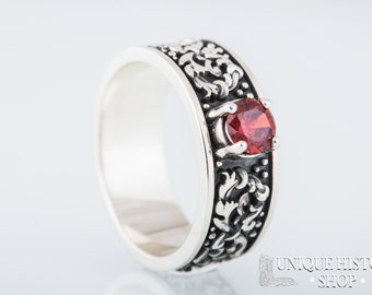 Puede incluir: Anillo de plata con una banda ornamentada y oscura con una piedra preciosa roja. El anillo tiene un patrón grabado detallado y un acabado pulido. La piedra preciosa es redonda y está engastada en un bisel elevado.