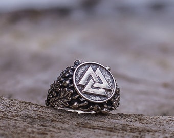 May include: A silver ring with a detailed design featuring a circular center with a raised, stylized triangular symbol. The ring is set on a piece of weathered wood.