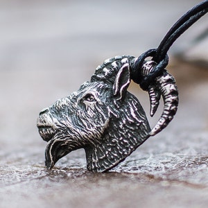 May include: Silver pendant necklace featuring a detailed goat head with curled horns. The pendant is attached to a black leather cord.