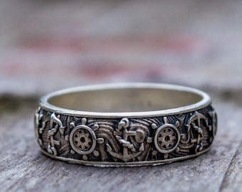 May include: A silver ring with a detailed design of anchors, ship wheels, and waves. The ring is on a wooden surface.