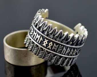 May include: Two silver rings, one with a band of stylized feathers and runes, the other a plain band. The ring with feathers and runes is on top of the plain band.