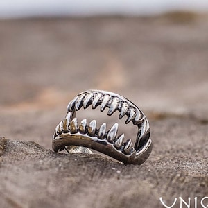 May include: A silver ring shaped like a shark's jaw, with sharp teeth. The ring is set against a blurred background and a wooden surface. The ring is a unique piece of jewelry.