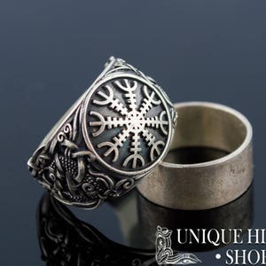 May include: A silver ring with a detailed design featuring a circular symbol with eight spokes. The ring is on a black surface and is reflected in the surface.