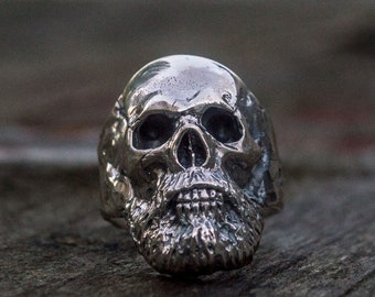 May include: A silver skull ring with a detailed beard. The ring is on a wooden surface.